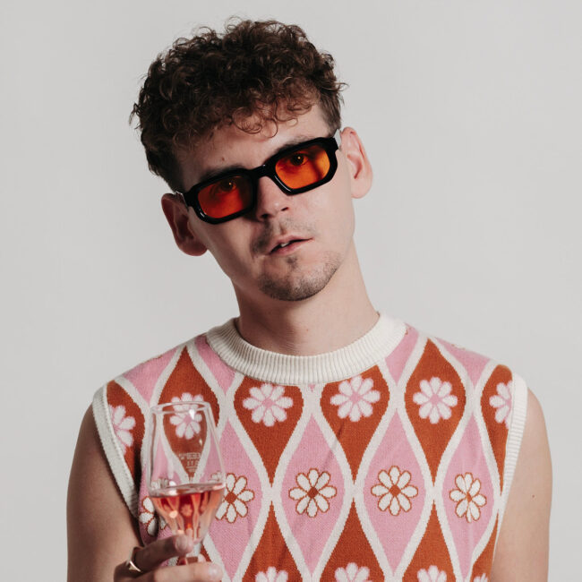 Michael Steiger trägt eine farblich abgestimmte getönte Brille zum floral gemusterten Polunder und hält ein Rosé Glas in der Hand.